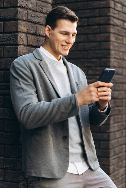 Современный красивый улыбающийся мужчина, стоящий у стены на улице с телефоном - Фото, изображение