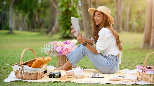 リアビュー、魅力的なアジアの若い女性が庭で美しいピクニックをしている間にタブレットでオンライン電子書籍を読む. - 写真・画像