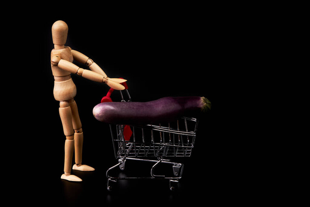 Dummy pushing a shopping cart with Japanese eggplant on black background. Trade symbol. Healthy shopping - Фото, изображение