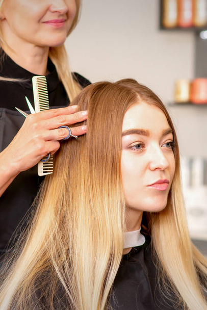 Gorgeous blonde long hair woman and her hairdresser looking and checking out hairstyle. Beautiful hairstyle of a young blonde woman after dying hair in a beauty salon, results of hair treatment - Zdjęcie, obraz