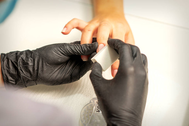 El primer plano de las manos caucásicas de una manicurista profesional está archivando las uñas de una mujer joven. Mujer caucásica joven recibiendo una manicura por una esteticista con una lima de uñas en un salón de uñas - Foto, imagen