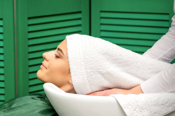 Peluquería envuelve el cabello de una joven mujer caucásica en una toalla blanca después de lavar la cabeza en el salón de peluquería - Foto, imagen