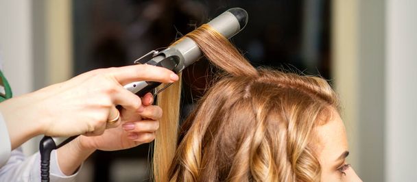 Kampaaja tekee kiharat curling-raudalla nuorelle naiselle, jolla on pitkät ruskeat hiukset kauneushoitolassa - Valokuva, kuva