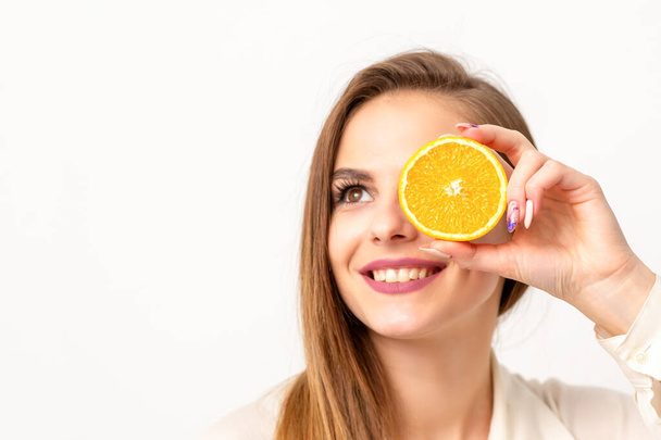 Retrato de uma jovem caucasiana alegre cobrindo o olho com uma fatia de laranja vestindo uma camisa branca sobre um fundo branco - Foto, Imagem