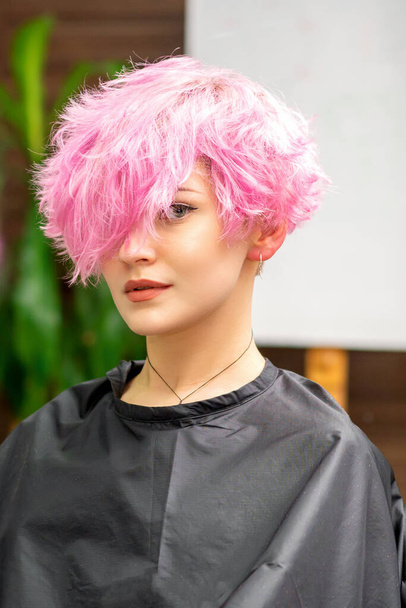 La hermosa joven mujer caucásica con un nuevo peinado corto rosa en el reflejo del espejo mirando a la cámara en un salón de peluquería - Foto, Imagen