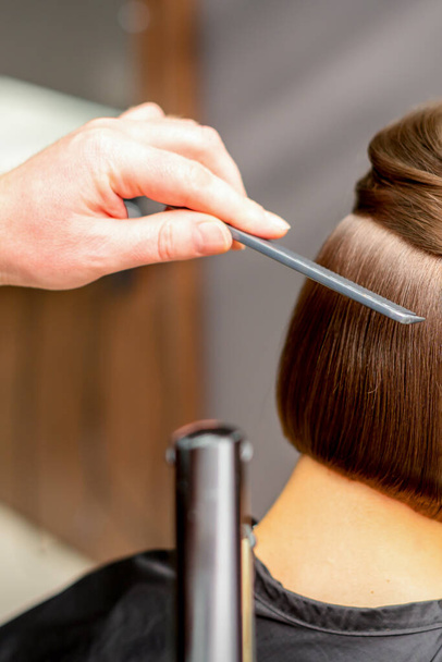 Парикмахер выпрямляет короткие волосы молодой брюнетки с плоским утюгом в парикмахерской, крупным планом - Фото, изображение