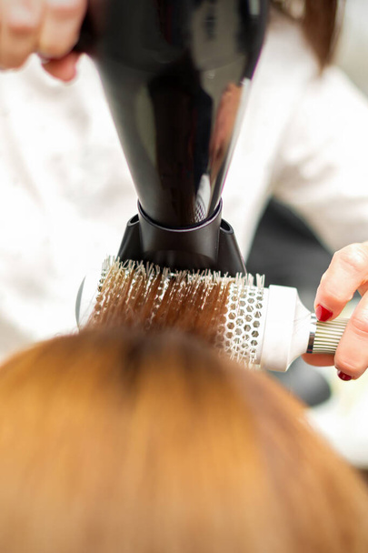 Een professionele kapper droogt lang rood haar met een haardroger en ronde borstel, close-up - Foto, afbeelding