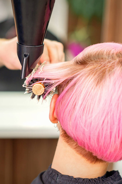 Ένας επαγγελματίας κομμωτής στεγνώνει μακριά κόκκινα μαλλιά με στεγνωτήρα μαλλιών και στρογγυλό πινέλο, κοντά - Φωτογραφία, εικόνα