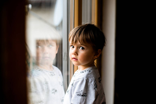 Sad kid looking out the window. Reflection in window glass. Face of two years old boy. Alone child. Little migrant child cry. Portrait of kid. Evacuation children. War Ukraine. Refugee abroad - Photo, Image