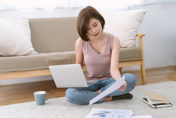 Портрет красивої молодої азіатської жінки перевіряє дані на ноутбуці і тримає документи, сидячи на підлозі з документами поруч з нею. Робота вдома, нова нормальна думка. - Фото, зображення