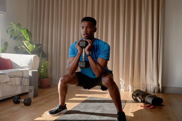 Junger afroamerikanischer Mann trainiert konzentriert von zu Hause aus, während er mit einer Hantel im Wohnzimmer hockt und zu Hause trainiert - Foto, Bild