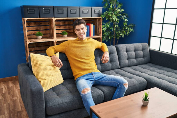 Fiatal spanyol férfi mosolyog magabiztos ül kanapén otthon - Fotó, kép