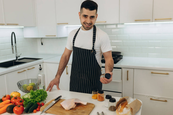 Portrait of a handsome man in the kitchen. The man is preparing food. Dressed in a black apron. Holds spices in his hands. The man looks at the camera and smiles - Photo, image