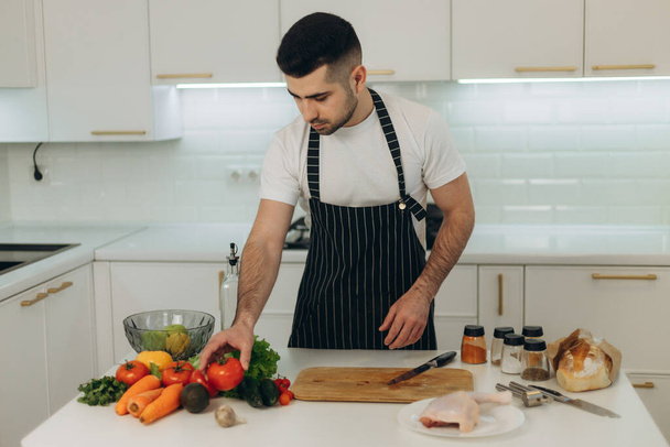 Porträt eines gutaussehenden Mannes in der Küche. Ein Mann in schwarzer Schürze. Gemüse zum Kochen hacken - Foto, Bild