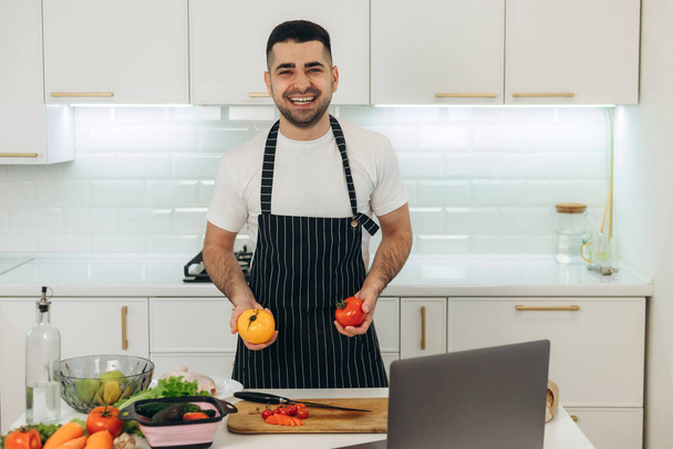 Glimlachende man bereidt groenten in de keuken gekleed in een zwart schort - Foto, afbeelding