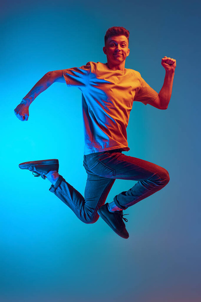 Dynamisches Porträt eines jungen, aufgeregten Mannes, der vor Aufregung fröhlich über blauen Studiohintergrund springt. Action, Sport, Mode - Foto, Bild