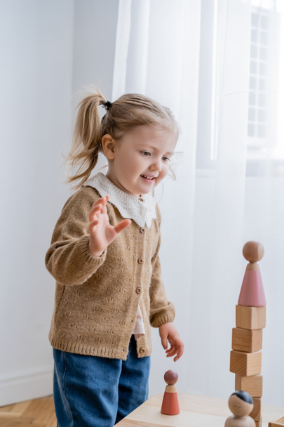 happy girl looking at tower made of wooden cubes and figurine while playing at home - Foto, Bild
