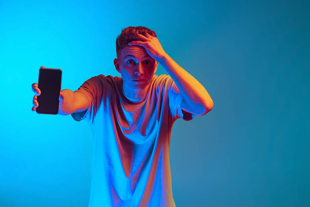 Studio shot of young emotional man, student using smarphone, showing phone screen isolated over blue studio background. Action, sales, fashion - Фото, зображення