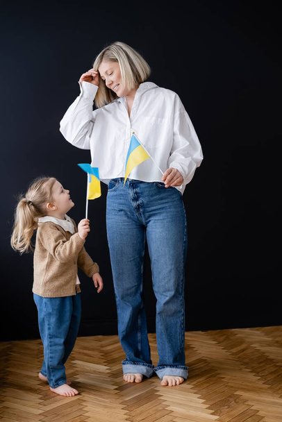 volledige weergave van blootsvoets moeder en dochter met kleine Oekraïense vlaggen glimlachend naar elkaar in de buurt van zwarte muur - Foto, afbeelding