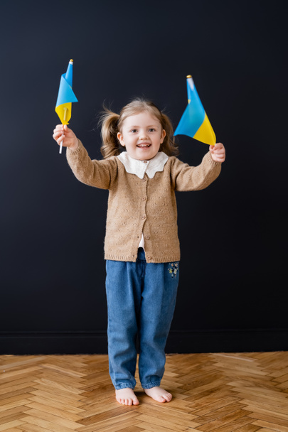 vista completa de niño patriótico alegre sosteniendo banderas ucranianas cerca de la pared negra - Foto, imagen