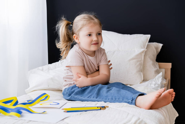 barefoot girl sitting with crossed arms near blue and yellow ribbon and color pencils on bed - Photo, Image