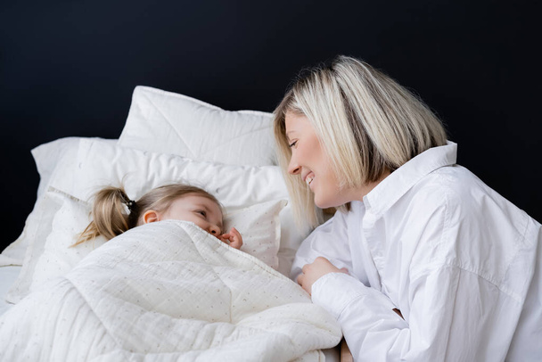 blonde woman smiling near little daughter lying under white blanket - Zdjęcie, obraz