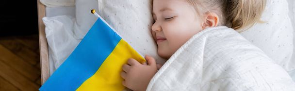 top view of smiling girl sleeping with small ukrainian flag in bed, banner - Photo, Image