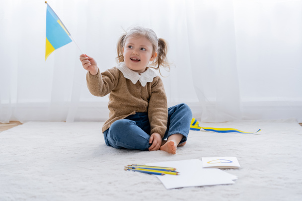 happy girl with small ukrainian flag sitting on floor near color pencils and card with blue and yellow heart - Photo, Image