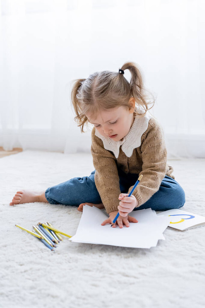 маленька дівчинка з хвостами малює на підлозі біля картки з синьо-жовтим серцем
 - Фото, зображення
