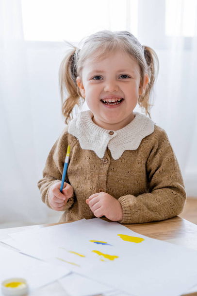 cheerful girl holding paintbrush near papers with blue and yellow spills - Fotografie, Obrázek