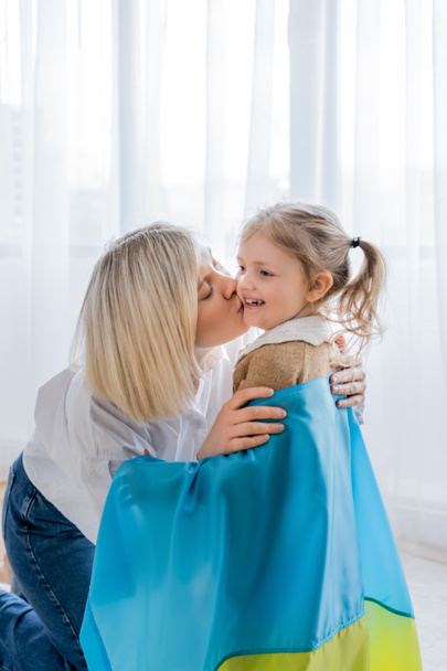blonde woman kissing cheerful daughter covered with ukrainian flag - Valokuva, kuva