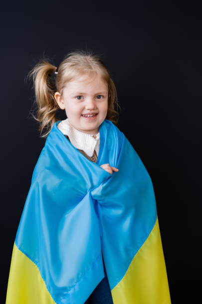 Ευτυχισμένο κορίτσι καλυμμένο με ουκρανική σημαία κοιτάζοντας κάμερα απομονωμένη σε μαύρο - Φωτογραφία, εικόνα