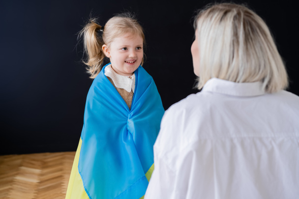 Маленька дівчинка, вкрита прапором українки, посміхається вдома біля розмитої мами. - Фото, зображення