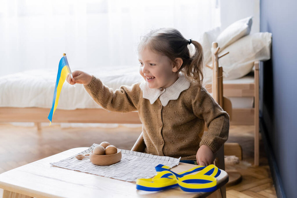 niña feliz sosteniendo bandera ucraniana cerca de cinta azul y amarillo y juguetes de madera en el escritorio - Foto, Imagen