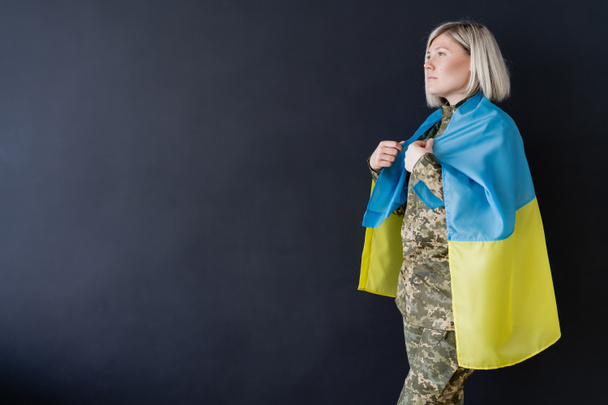 πλευρική άποψη της στρατιωτικής γυναίκας με ουκρανική σημαία στους ώμους κοιτάζοντας μακριά απομονώνονται σε μαύρο - Φωτογραφία, εικόνα