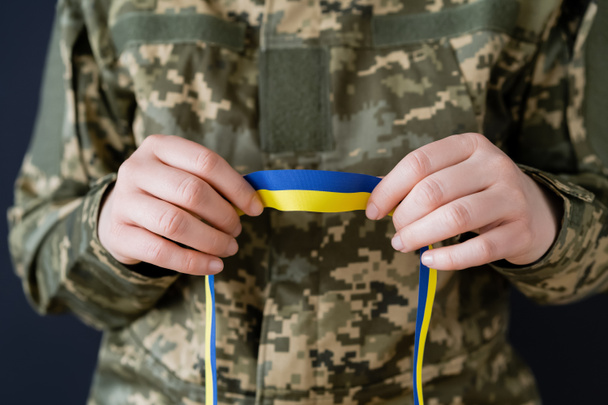 vista parcial de la mujer en uniforme militar sosteniendo cinta azul y amarilla aislada en negro - Foto, Imagen