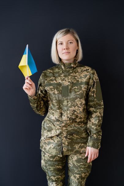 Frontansicht einer Militärfrau mit ukrainischer Flagge, die isoliert auf schwarz in die Kamera blickt - Foto, Bild