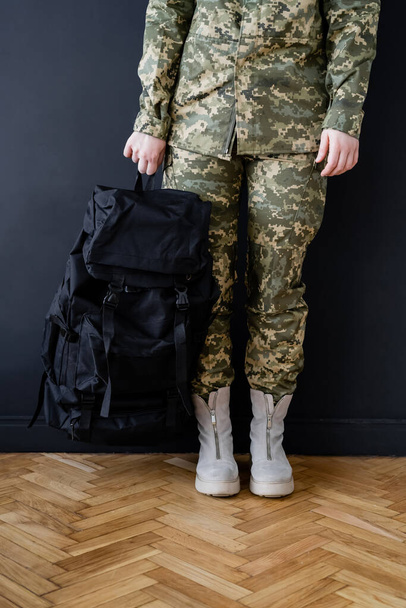 cropped view of military woman with black backpack near dark wall - Foto, Imagem