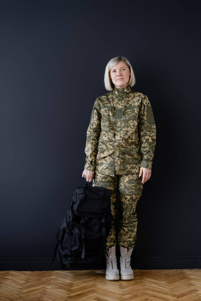 longitud completa de la mujer en uniforme militar de pie con mochila negra cerca de la pared oscura - Foto, imagen