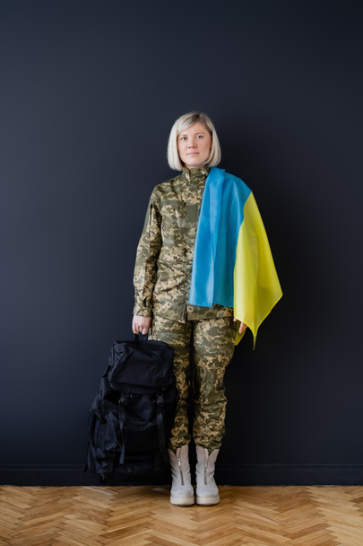 longitud completa de la mujer militar con bandera ucraniana y mochila cerca de la pared negra - Foto, Imagen