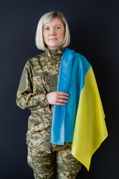 rubia militar con bandera ucraniana mirando a la cámara aislada en negro - Foto, Imagen