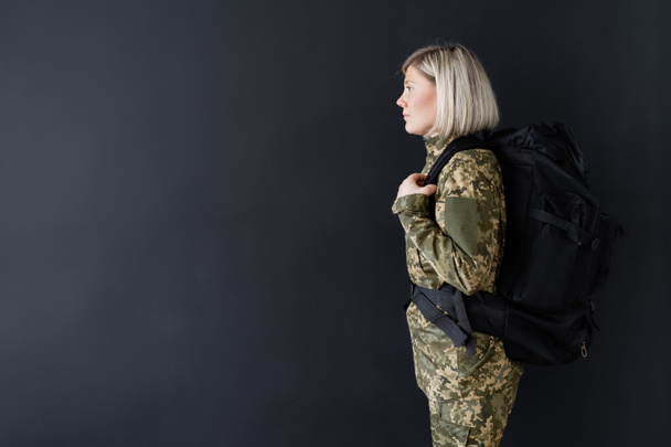 zijaanzicht van blond militair vrouw met rugzak geïsoleerd op zwart - Foto, afbeelding