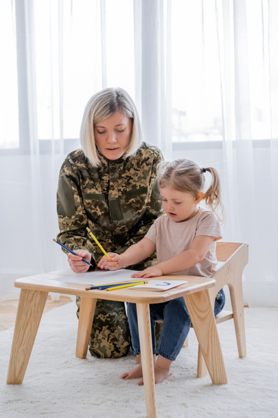 blonde military woman and little daughter drawing with blue and yellow pencils at home - Photo, Image