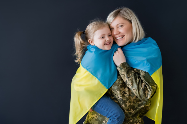 feliz militar mujer e hija cubierta con bandera ucraniana abrazo aislado en negro - Foto, Imagen