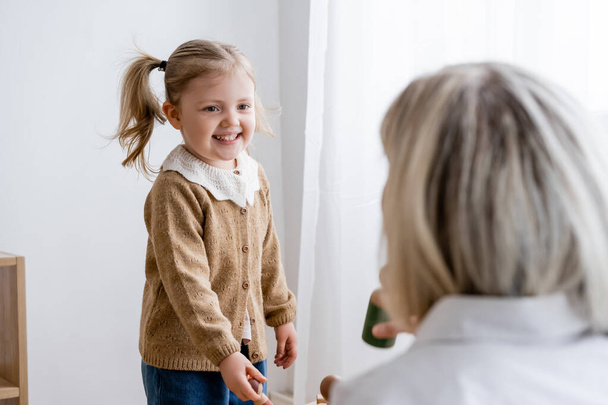 cheerful girl with ponytail smiling near blurred mom at home - Foto, immagini