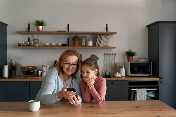 Caucasian grandmother and her granddaughter using smart phone together and sitting in the kitchen - Fotografie, Obrázek