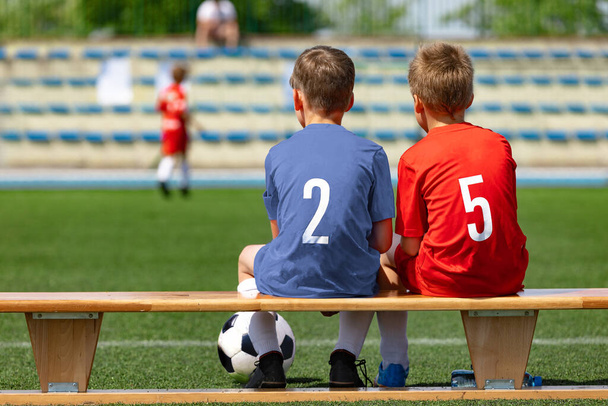 Due amici sportivi maschili seduti su una panchina di legno a guardare una partita di calcio. Ragazzi della scuola in uniforme da calcio. Bambini felici che praticano sport all'aperto. Amicizia tra due squadre sportive opposte - Foto, immagini