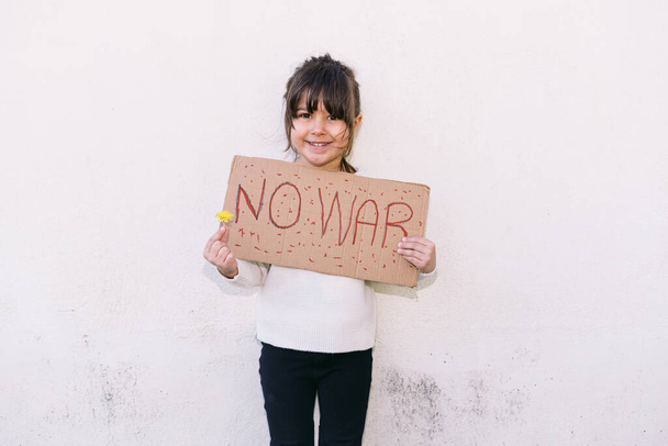 Mała dziewczynka aktywistka uśmiechnięta, przeciwko wojnie trzyma napis "NIE WOJNA" i żółty kwiat. Koncepcja wojny, konfrontacji, aktywizmu, pokoju i miłości. - Zdjęcie, obraz
