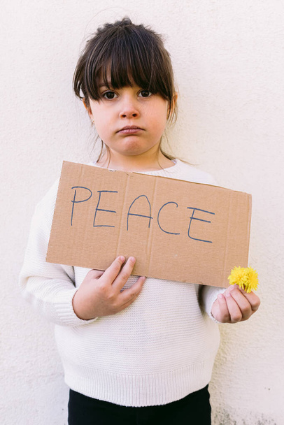 真剣に、反戦活動家の少女は、「平和」と黄色の花という看板を掲げている。戦争、対立、行動主義、平和と愛の概念. - 写真・画像