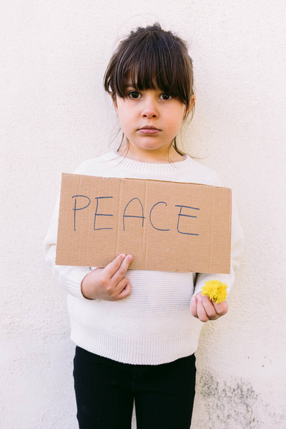 Σοβαρό, αντιπολεμικό ακτιβιστικό κοριτσάκι που κρατά μια πινακίδα που γράφει: "ΕΙΡΗΝΗ" και ένα κίτρινο λουλούδι. Έννοια του πολέμου, αντιπαράθεση, ακτιβισμός, ειρήνη και αγάπη. - Φωτογραφία, εικόνα